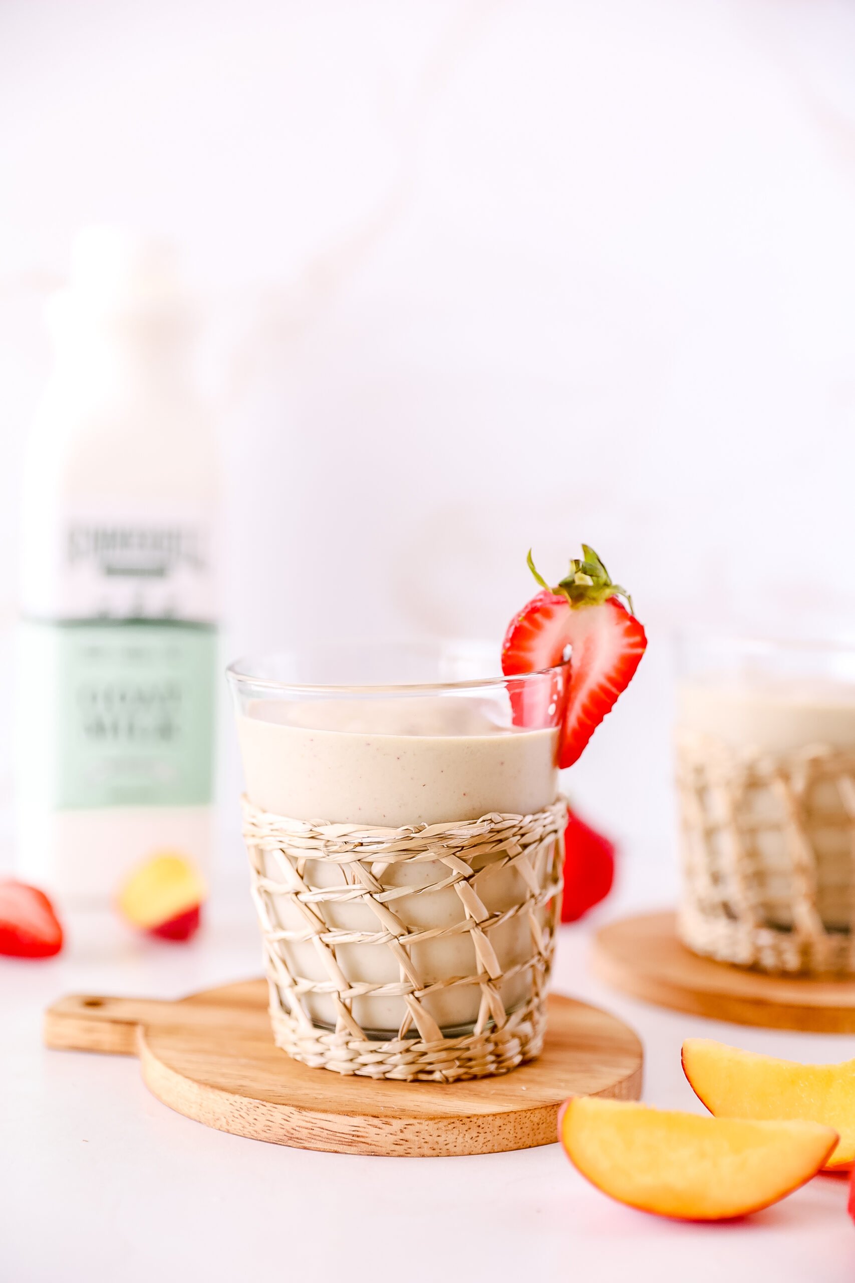 A glass of peach smoothie sits on a small round cutting board, with a wicker wrap and strawberry slice for garnish. A bottle of Summerhill goat milk sits in the left background. peach slices and strawberries are scattered on the counter for decoration.