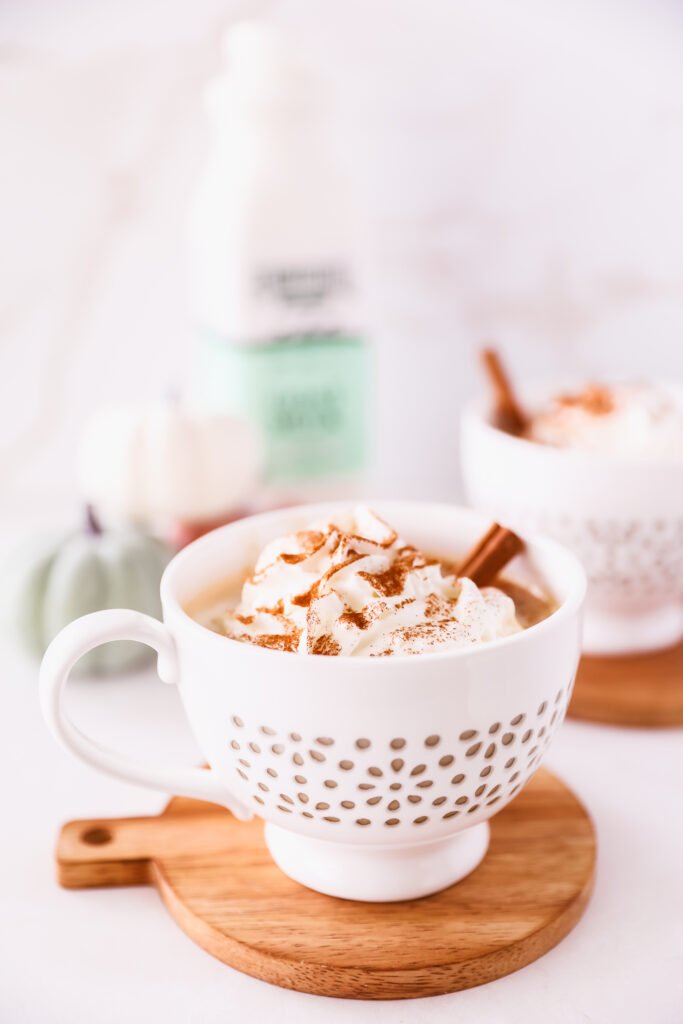 A white mug full of coffee, whipped cream, cinnamon, and a cinnamon stick sit on a small circular wooden cutting board. A blurred bottle of Summerhill Goat Milk and a second latte sit in the background
